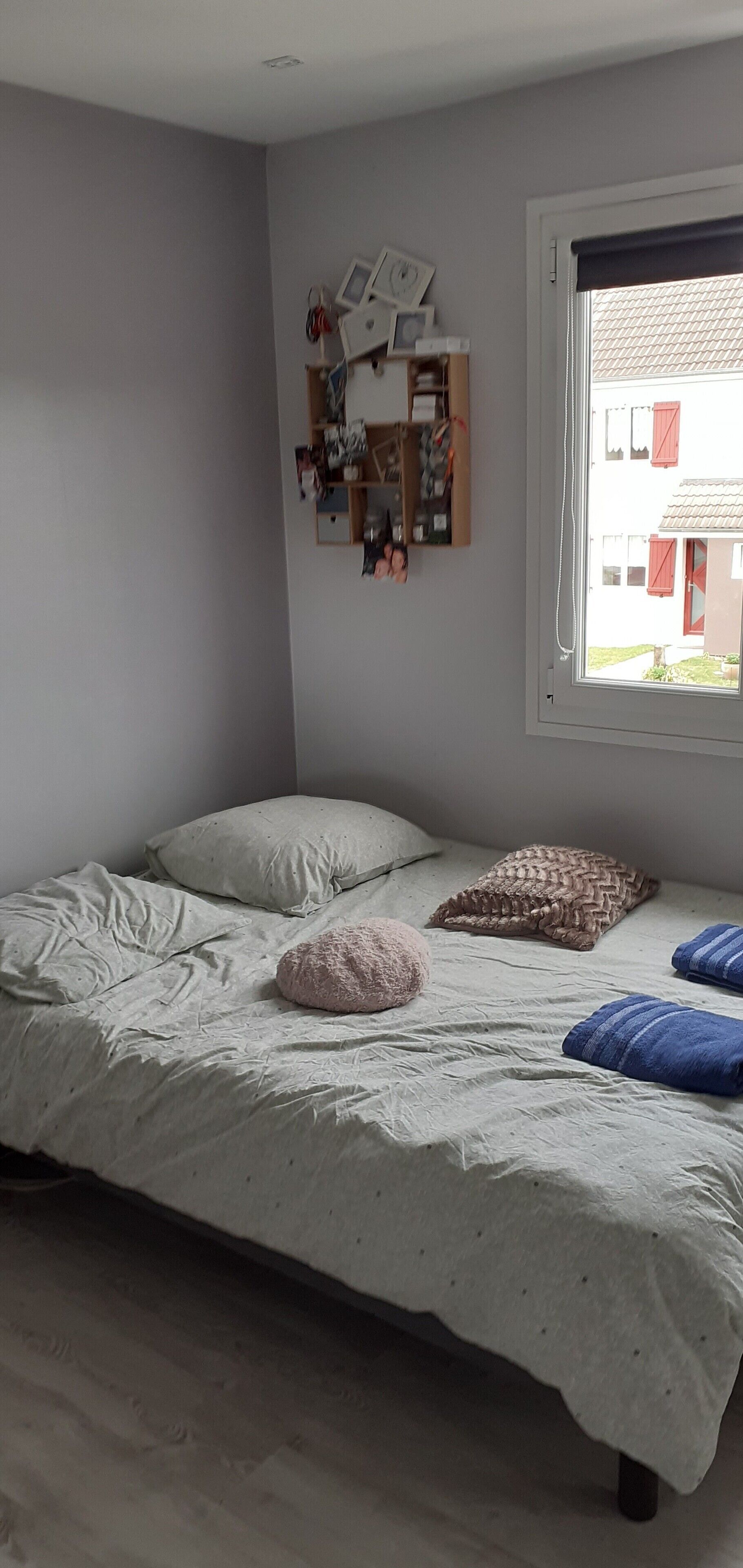 A bedroom with a bed, pillows, a bedside table with a lamp, a window with blinds, and a wall-mounted shelf with various items.