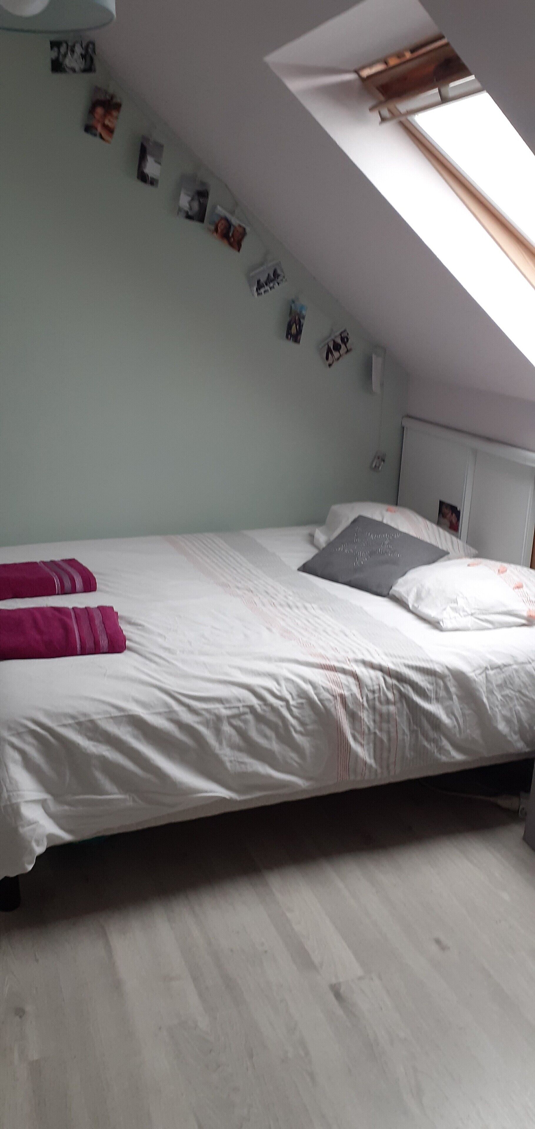 Een slaapkamer met dakraam, een bed met witte beddengoed en een roze deken, en verschillende foto's aan de muur.