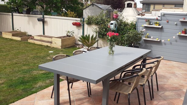 A patio with a grey table and wicker chairs, surrounded by a garden with raised beds and a small shed.