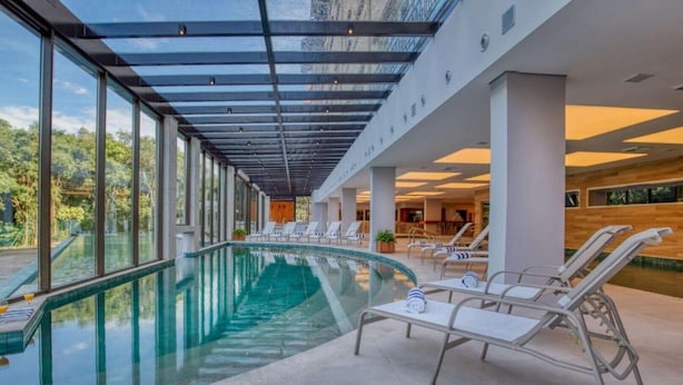 Una piscina interior moderna con paredes de vidrio transparente, sillones de descanso y vistas del paisaje circundante.