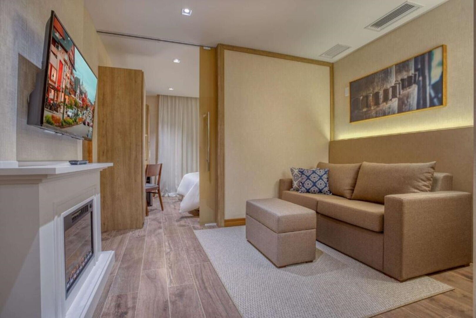 A modern hotel room with a sofa, ottoman, and a television mounted on the wall.