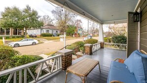 A covered porch with a wicker chair and a blue cushion, a brick wall, and a view of a street with houses and cars.