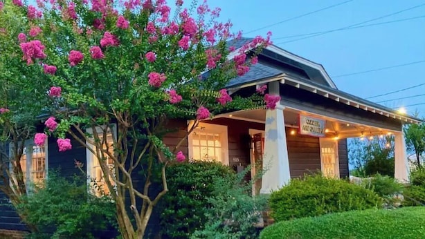 A wooden house with a porch, surrounded by lush greenery and flowering trees.