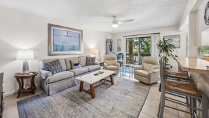 A spacious living room with a sofa, coffee table, and chairs, a dining area with a bar, and a large window with a view. - 2BR Forest Ridge condo with pool, balcony, WiFi, and beach access (Fernandina Beach)