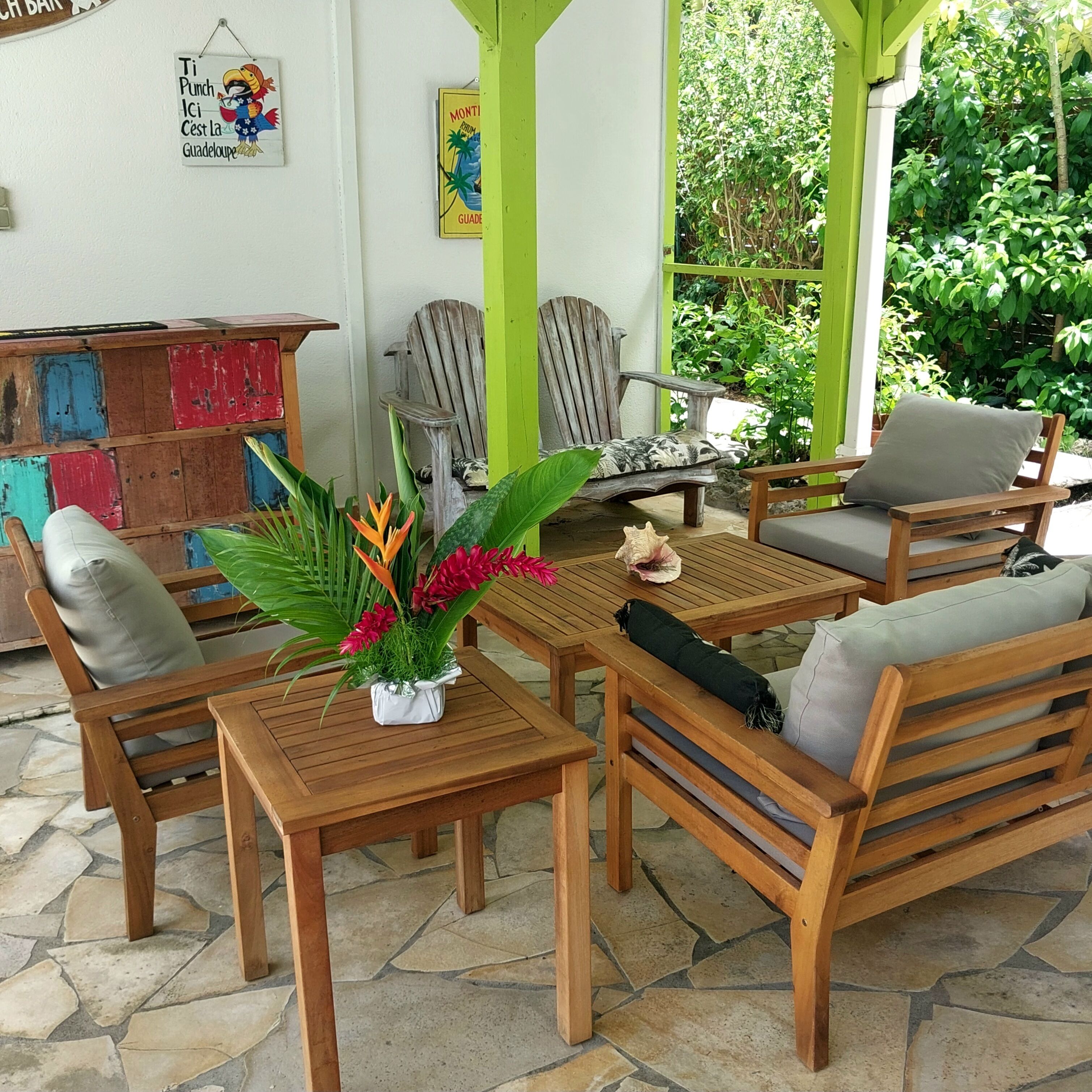 Un espacio de estar al aire libre con madera, que incluye una mesa y sillas, rodeado de follaje y un suelo de piedra.