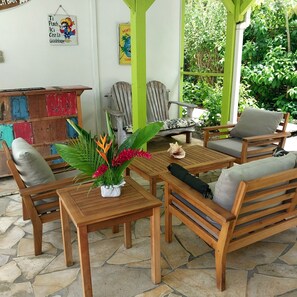 A wooden outdoor seating area with a table and chairs, surrounded by greenery and a stone floor.