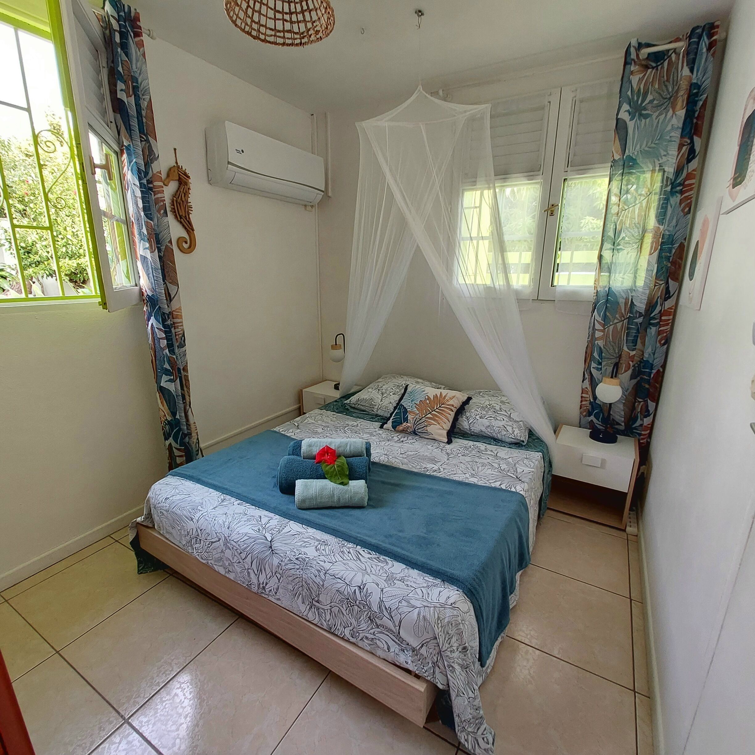 Un dormitorio con cama, mesita de noche, ventana con cortinas, aire acondicionado y mosquitera.