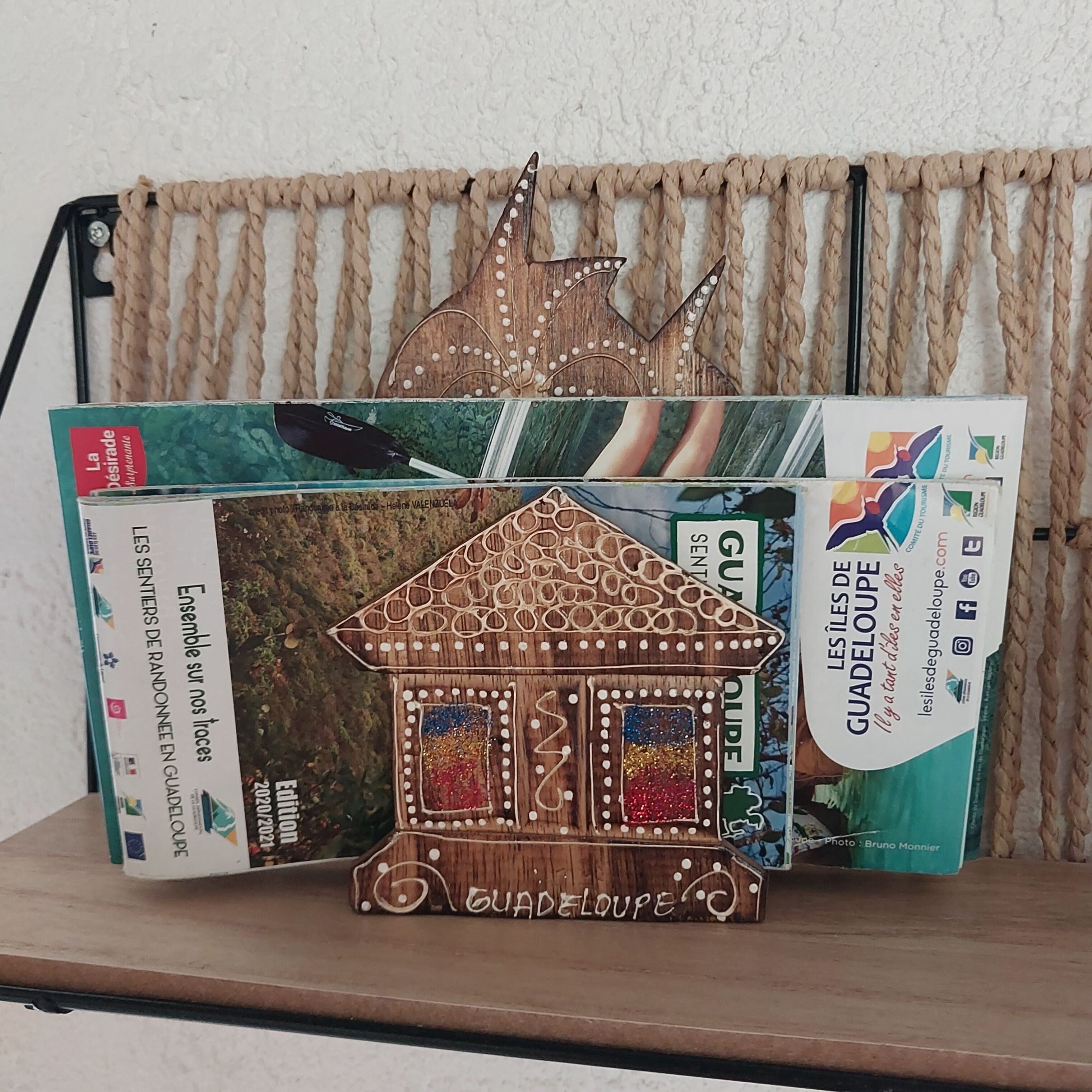 Une maquette en bois d’une maison de la Guadeloupe, avec un intérieur aux couleurs vives, placée sur une étagère devant un fond texturé.