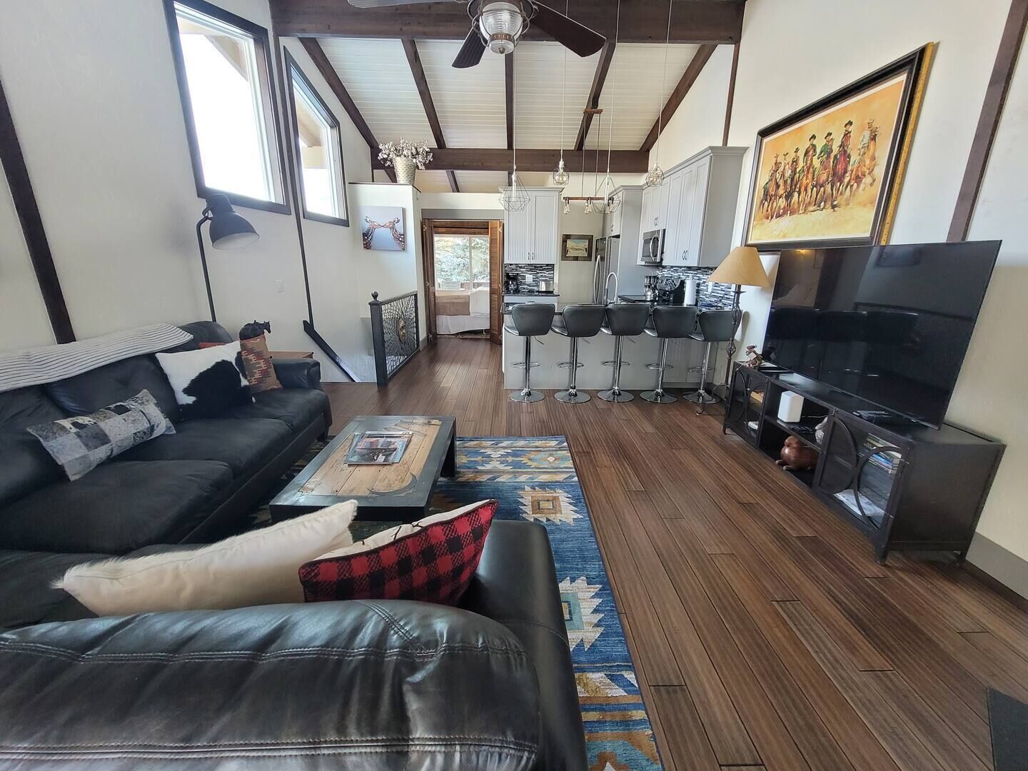 A modern living room with a black leather sofa, a coffee table, and a TV. There is a kitchen area with bar stools and a dining table.