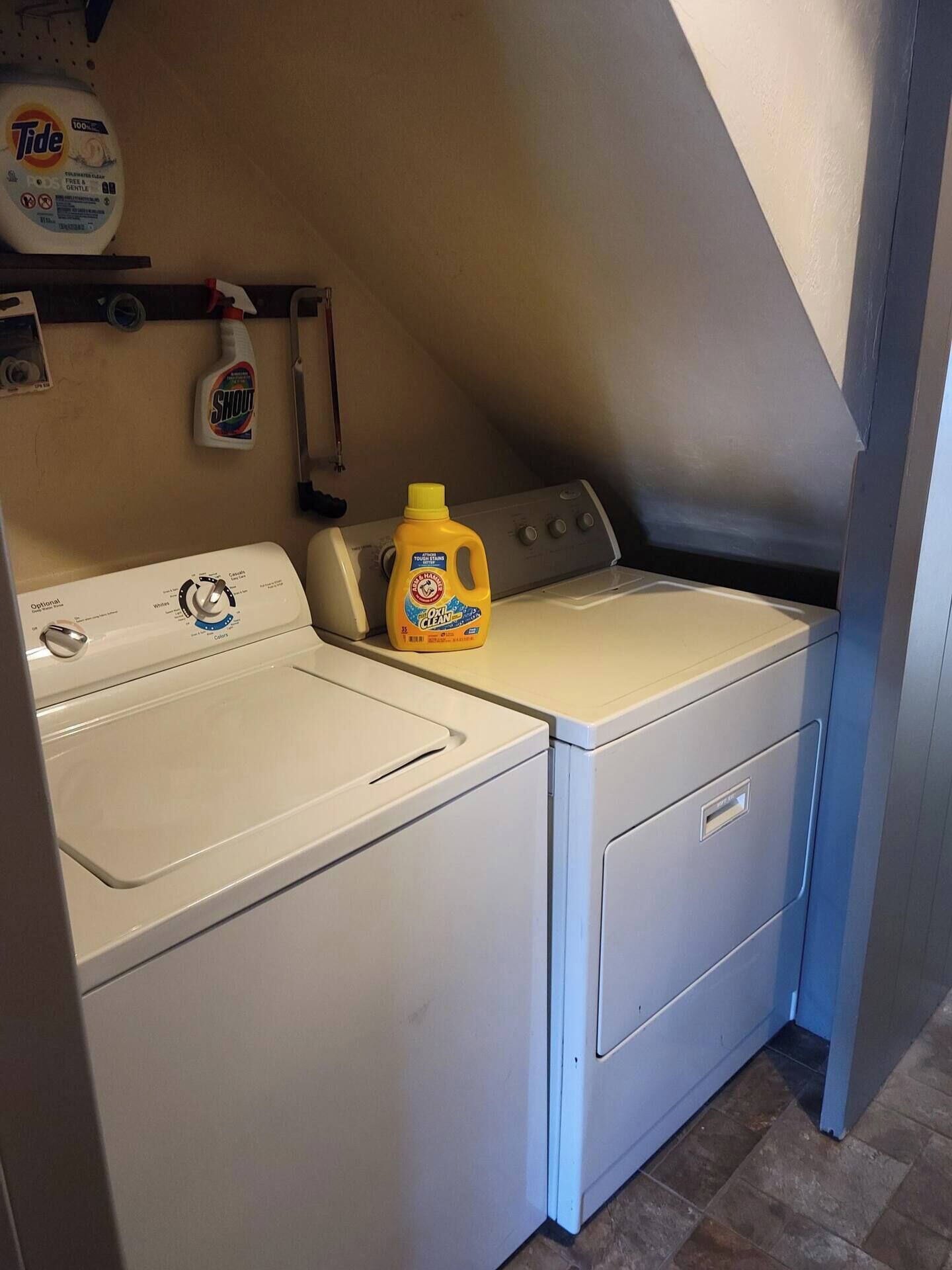 Een wasserette met twee witte wasmachines en drogers, een fles Tide wasmiddel en een fles Shout vlekverwijderaar op een plank.