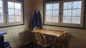 A wooden cabin interior with a dining table and chairs, two windows with curtains, and a coat hanging on the wall.