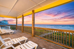 Terraza de madera con sillones de descanso con vista a una playa y al océano al atardecer.
