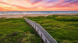 Een houten looppad dat naar een strand leidt, met een zonsondergang op de achtergrond.
