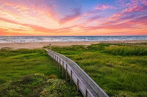 Una pasarela de madera que lleva a una playa, con un atardecer de fondo.