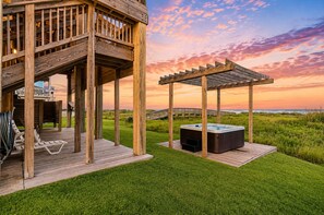 Una terraza de madera con jacuzzi y sillones de descanso, con vista a un espacio cubierto de césped, una pérgola de madera y el océano.