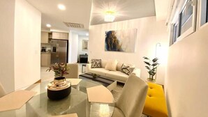 A modern living room with a glass coffee table, a sofa, and a yellow chair. There is a kitchen in the background with a refrigerator and a dining area. - Peninsula 12, 𖠁 3Bdr Lux Beach condo 𖠁Pools 𖠁Rooftop 𖠁Gym 𖠁 (Playa Langosta)