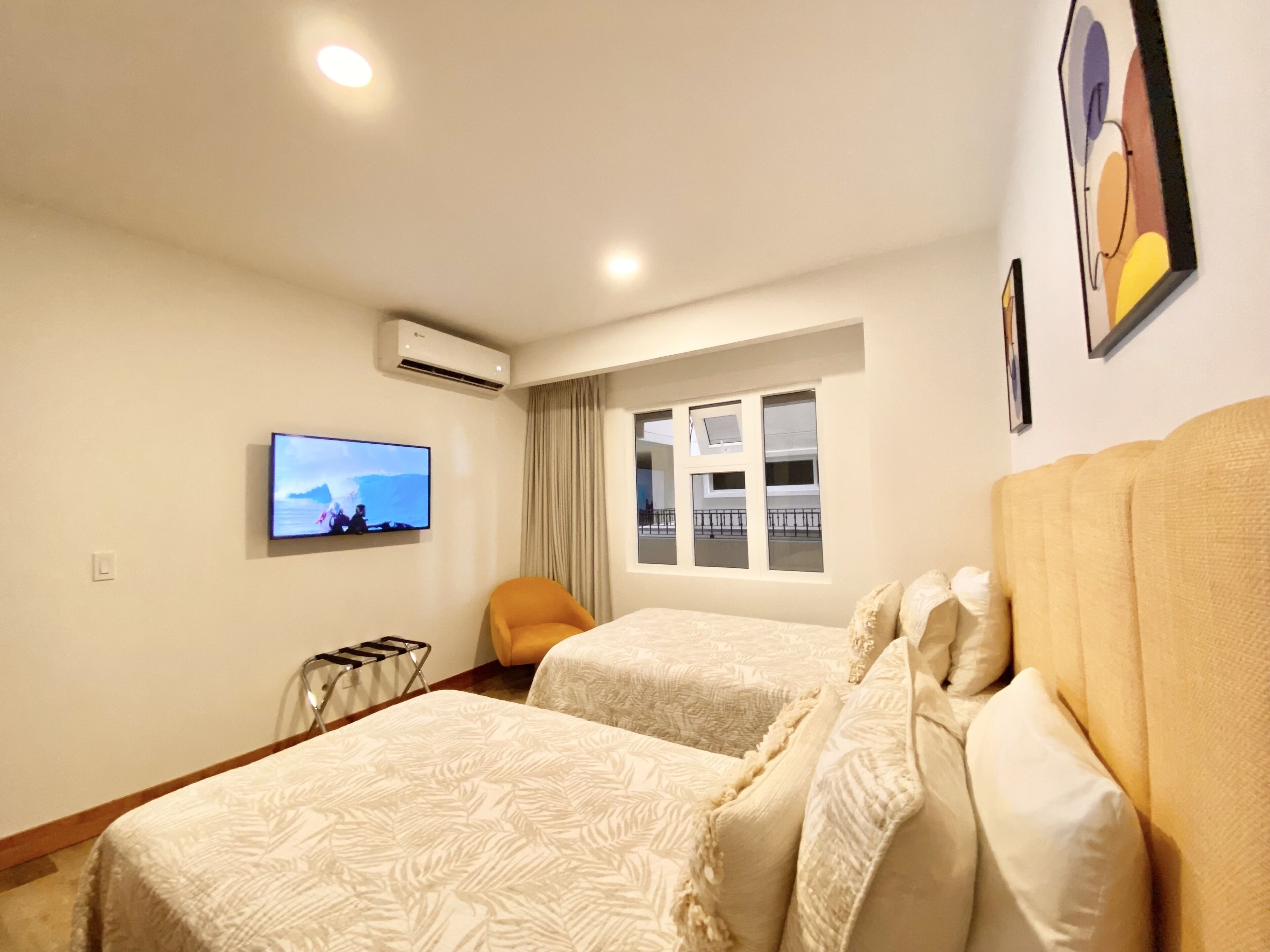 A hotel room with two beds, a TV, an air conditioner, a window with a view, and a chair.