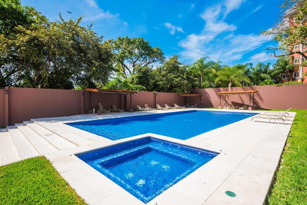 A rectangular swimming pool with a tiled deck, surrounded by a low wall and lush greenery.