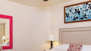 A hotel room with a bed, a ceiling fan, a nightstand with a lamp, a painting on the wall, and a mirror with a red frame.