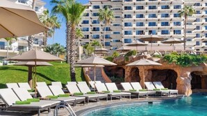 A hotel pool area with lounge chairs and umbrellas, surrounded by palm trees and a large building in the background.