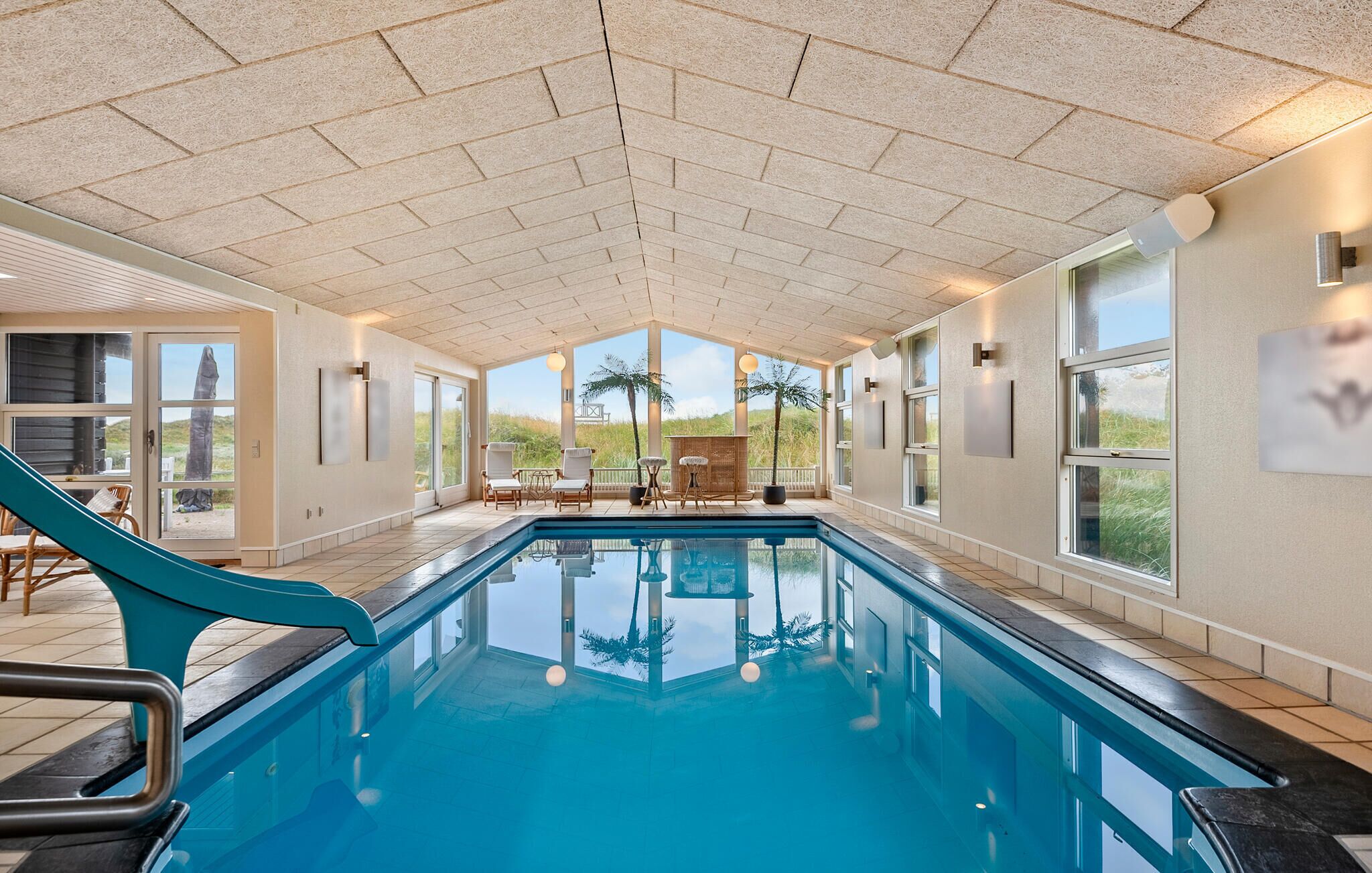 An indoor swimming pool with a slide, surrounded by large windows and a view of the outdoors.