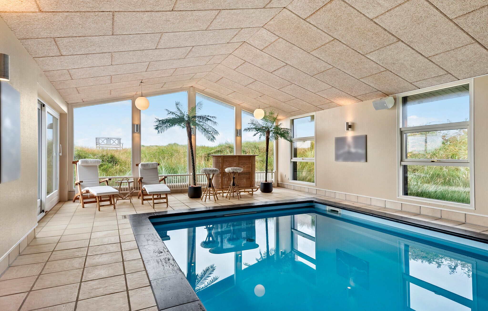 Un espace piscine intérieur moderne offrant une vue dégagée sur l’extérieur, notamment sur une plage et une pelouse.