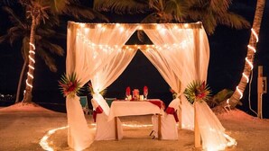 Un espace aménagé sur la plage avec une tente, une table et des chaises, décoré de guirlandes lumineuses et d’arrangements floraux.