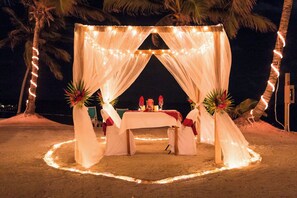 A beachside tent set up with a table and chairs, surrounded by palm trees and string lights.