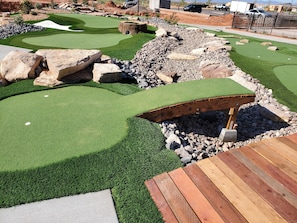 Een minigolfbaan met een houten brug, groene kunstmatige grasmat en omliggende rotsen.