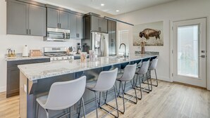 A modern kitchen with grey cabinets, a large island with bar stools, stainless steel appliances, and a bull painting on the wall. - New | 4 Bed | Heated Pool | Lazy River | Mini Golf | BI (Washington)