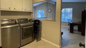 Uma lavanderia com máquina de lavar e secadora, um relógio na parede e vista para uma sala de estar com mesa de jantar e cadeiras.