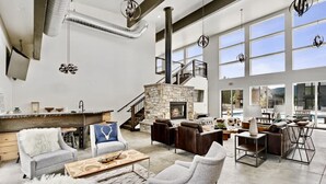 A spacious living area with a stone fireplace, leather sofas, and a high ceiling with exposed ductwork. - Private Sauna | Pool & Hot Tub | Awesome Views (Whitefish)