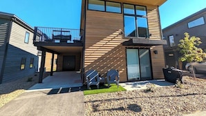 A modern two-story building with a balcony, a garage, and a small outdoor seating area. - Private Sauna | Pool & Hot Tub | Awesome Views (Whitefish)