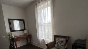 A room with a wooden desk, a chair with a cushion, a stone-patterned rug, a mirror on the wall, and a window with sheer curtains.
