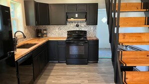 A modern kitchen with dark cabinetry, a wooden countertop, a black stove, and a wooden staircase. - The Archer House - 1 Block from Spring River.  2 Bedrooms + Loft Bedroom!   (Mammoth Spring)