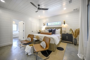 A bedroom with a bed, two chairs, a nightstand, a dresser, a ceiling fan, and a window.
