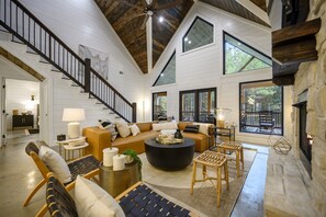 Amplia sala de estar con techo alto, escalera, chimenea y ventanas grandes.