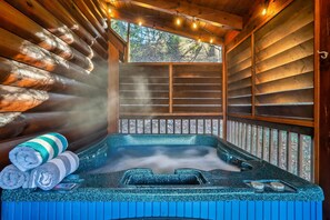 A hot tub with steam rising, surrounded by wooden walls and a window with a view of trees. - Cozy Cabin in the Hills. Hot tub, Views, Pool table, Pet friendly and Much More (Pigeon Forge)