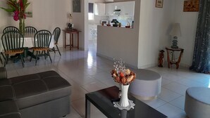 Ein Wohnzimmer mit einer Couch, einem Esstisch, Stühlen, einer Vase mit Blumen, einer Lampe und einer Küche im Hintergrund.