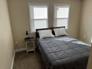 A bedroom with a bed, two windows, a nightstand, and a lamp.