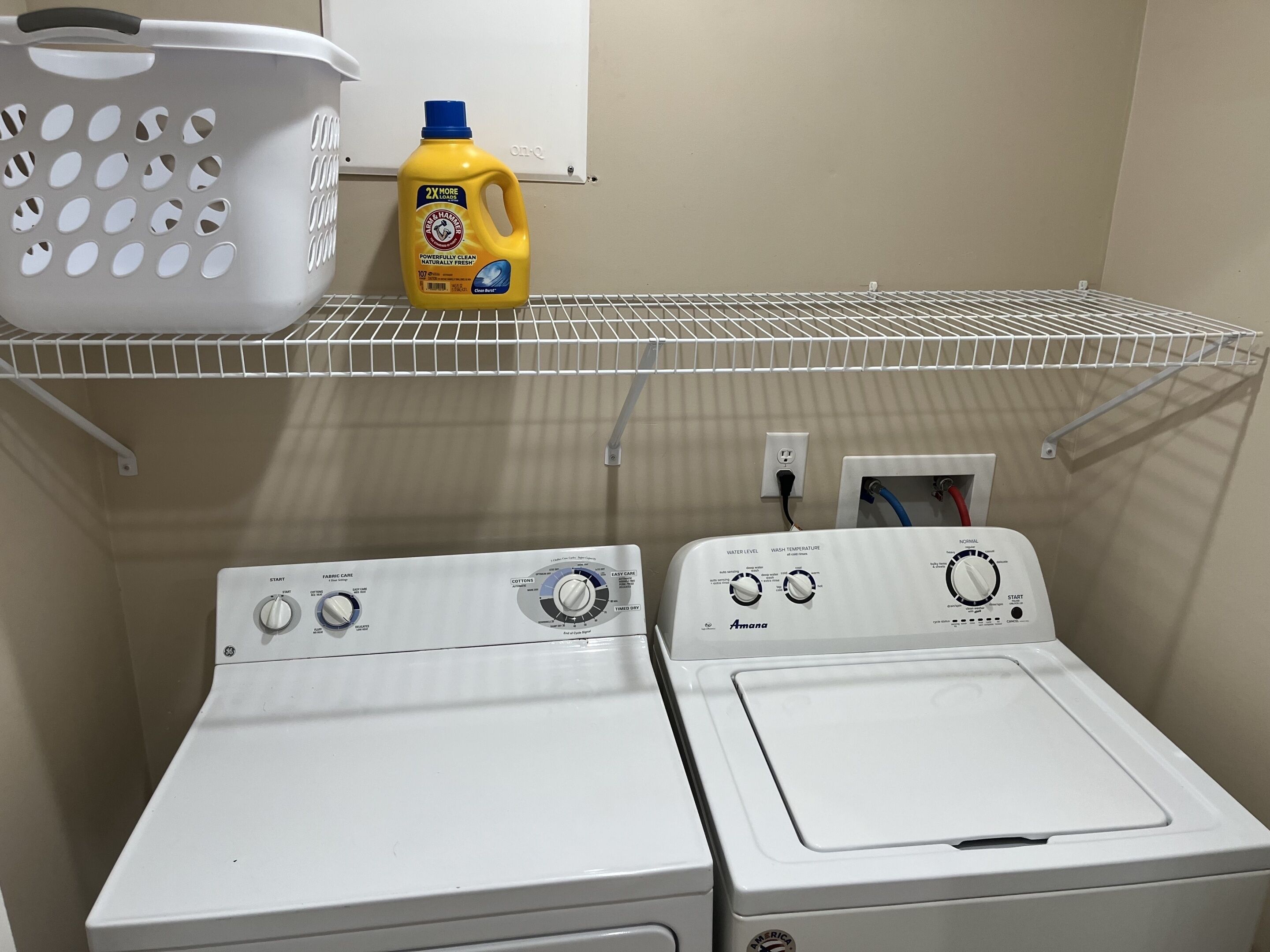 A laundry area with two white washing machines, a white basket, and a bottle of detergent on a shelf.