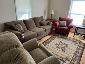 A living room with a brown sofa, a red armchair, a small table with a lamp, and a patterned rug.