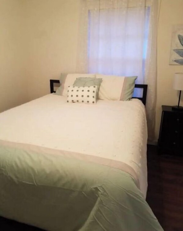 A neatly made bed with a white and green bedspread, a pillow with a polka dot pattern, and a plain wall with a window.