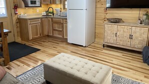 Eine kleine Küche mit Holzausstattung, einem weißen Kühlschrank, einer Mikrowelle und einem Toaster. Auf dem Boden liegt ein schwarzer Teppich und es gibt einen beigen Hocker im Sitzbereich.