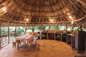Enjoy casual dining in this open-plan space with a thatched roof, wooden table, and kitchen area.