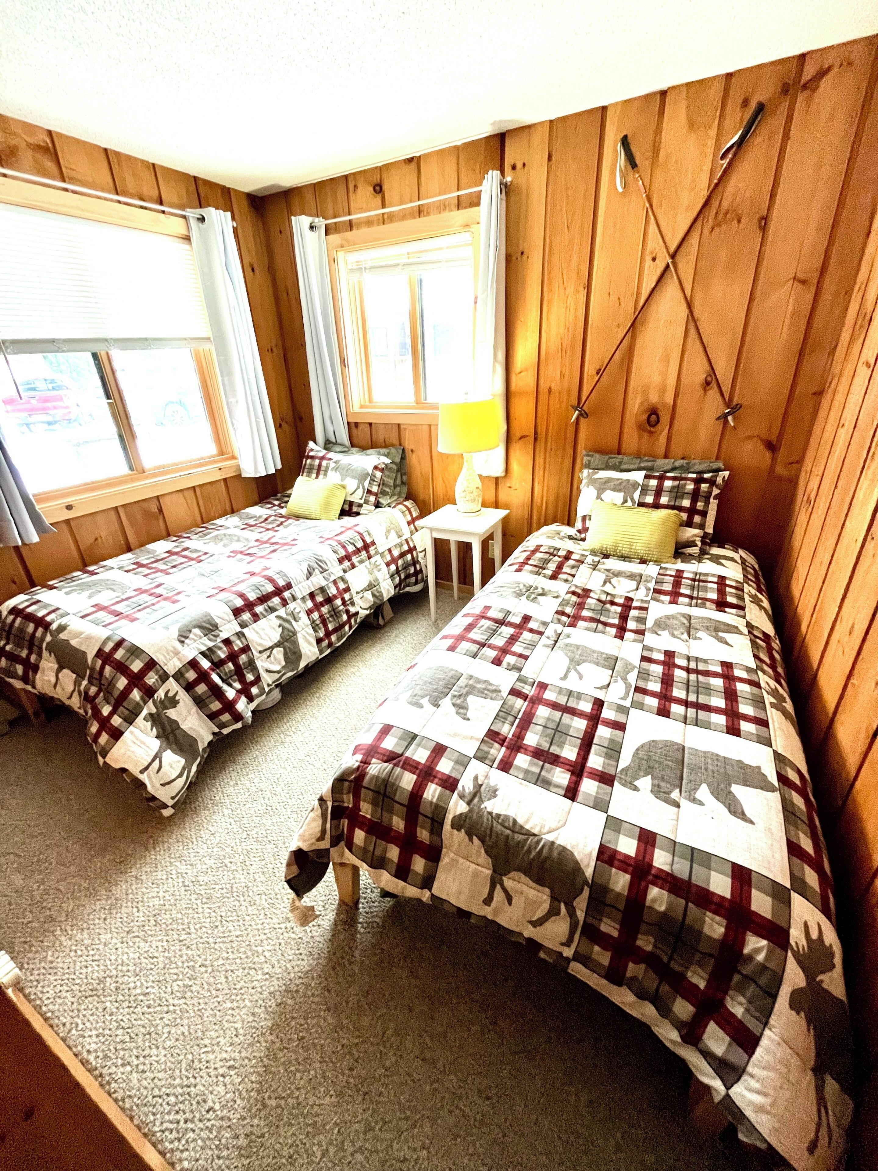 Ein Zimmer mit zwei Betten, Holzwänden, einem Fenster mit Vorhängen und einem kleinen Tisch mit einer Lampe.