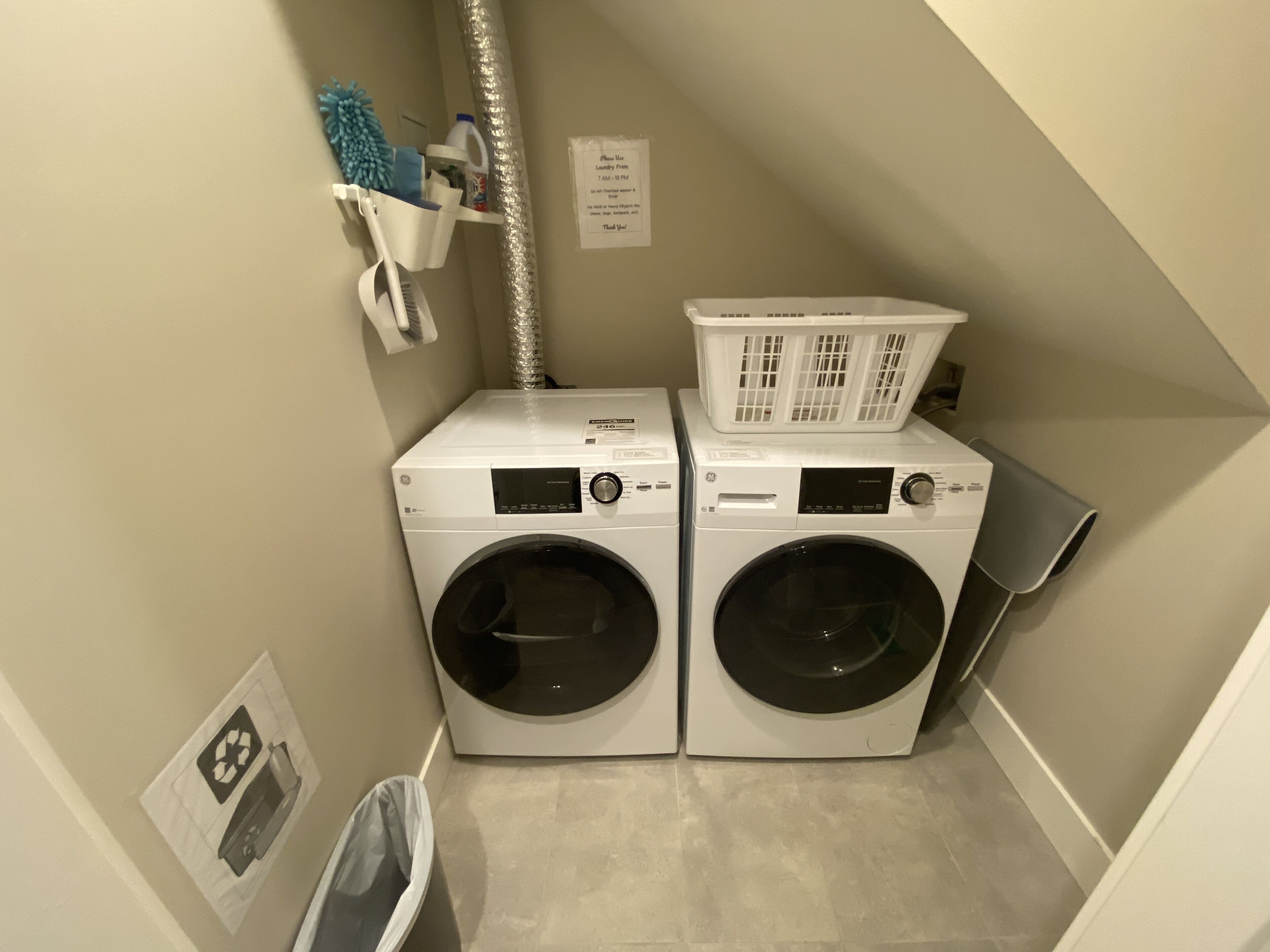 A laundry room with two front-loading washing machines, a drying rack, and a trash bin.