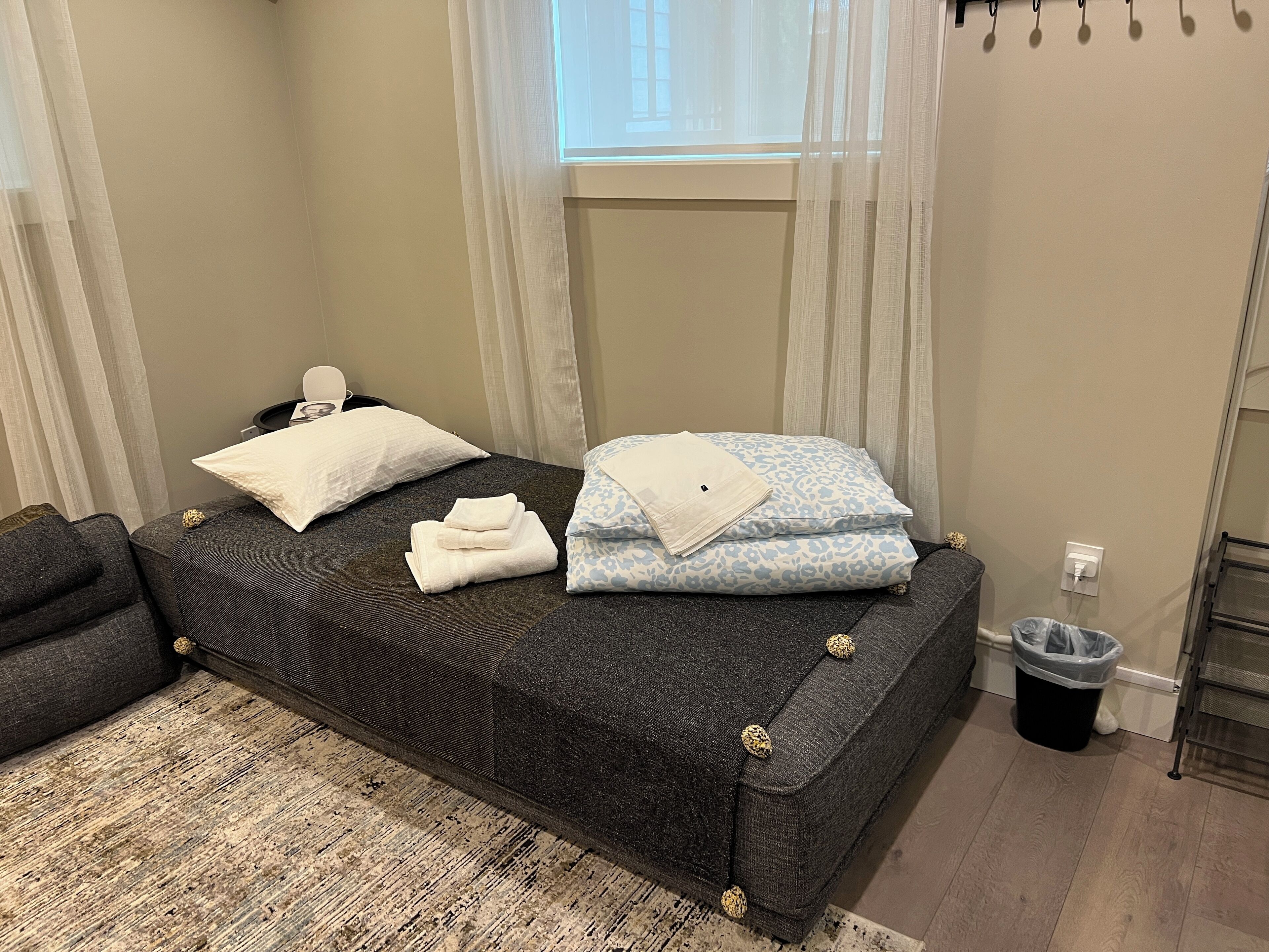 Una cama bien hecha con almohadas y toallas dobladas, una mesita de noche con lámpara, una ventana con cortinas ligeras y un cubo de basura.