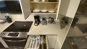 A modern kitchen with a built-in oven, a coffee maker, a toaster, and a kettle. There is a shelf with plates and bowls, and a drawer with utensils.