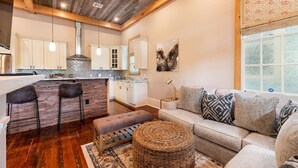 A modern kitchen with white cabinets, a wooden island, and bar stools. There is a living area with a sofa and ottoman, and a large window with patterned curtains. - "Life of Riley" 3bd/3ba Irish Channel Home! (New Orleans)
